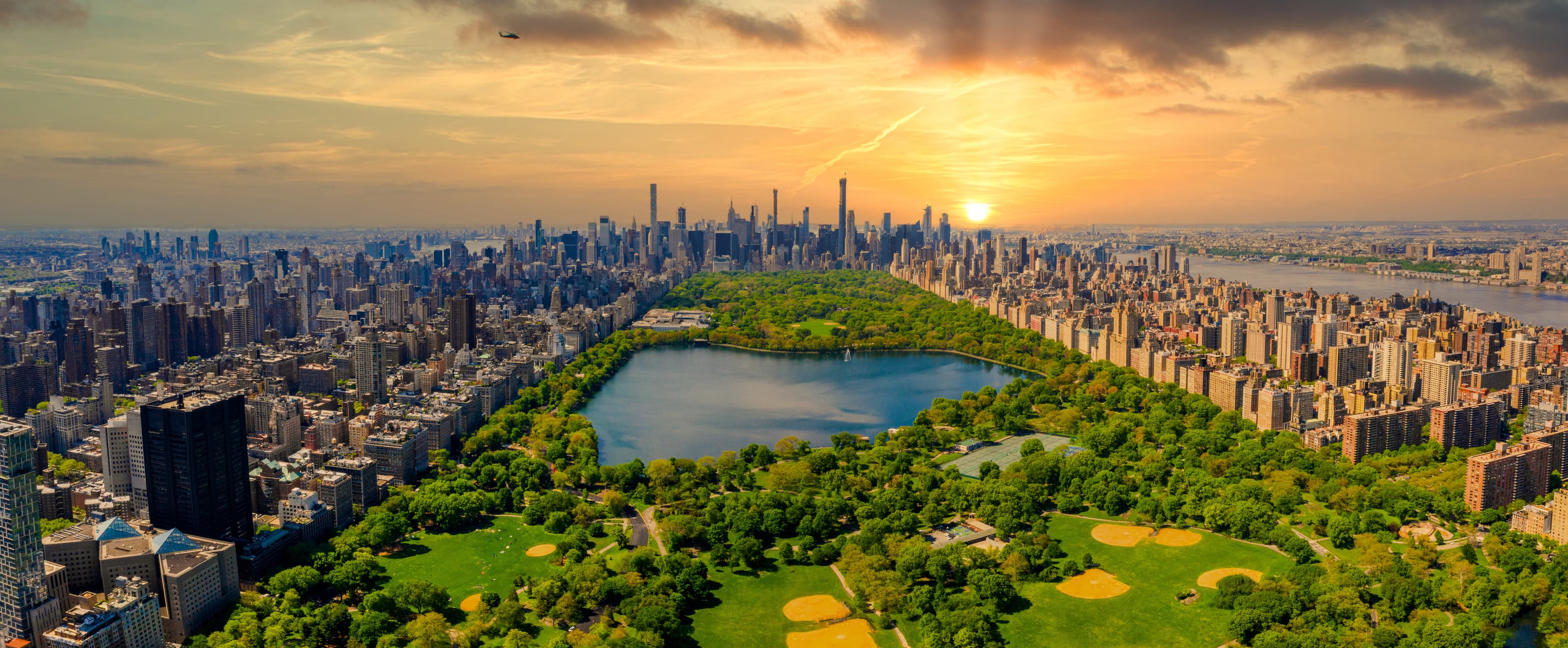 An aerial view of Central Park and the Manhattan skyline at golden hour, the reservoir glinting below a warm sky