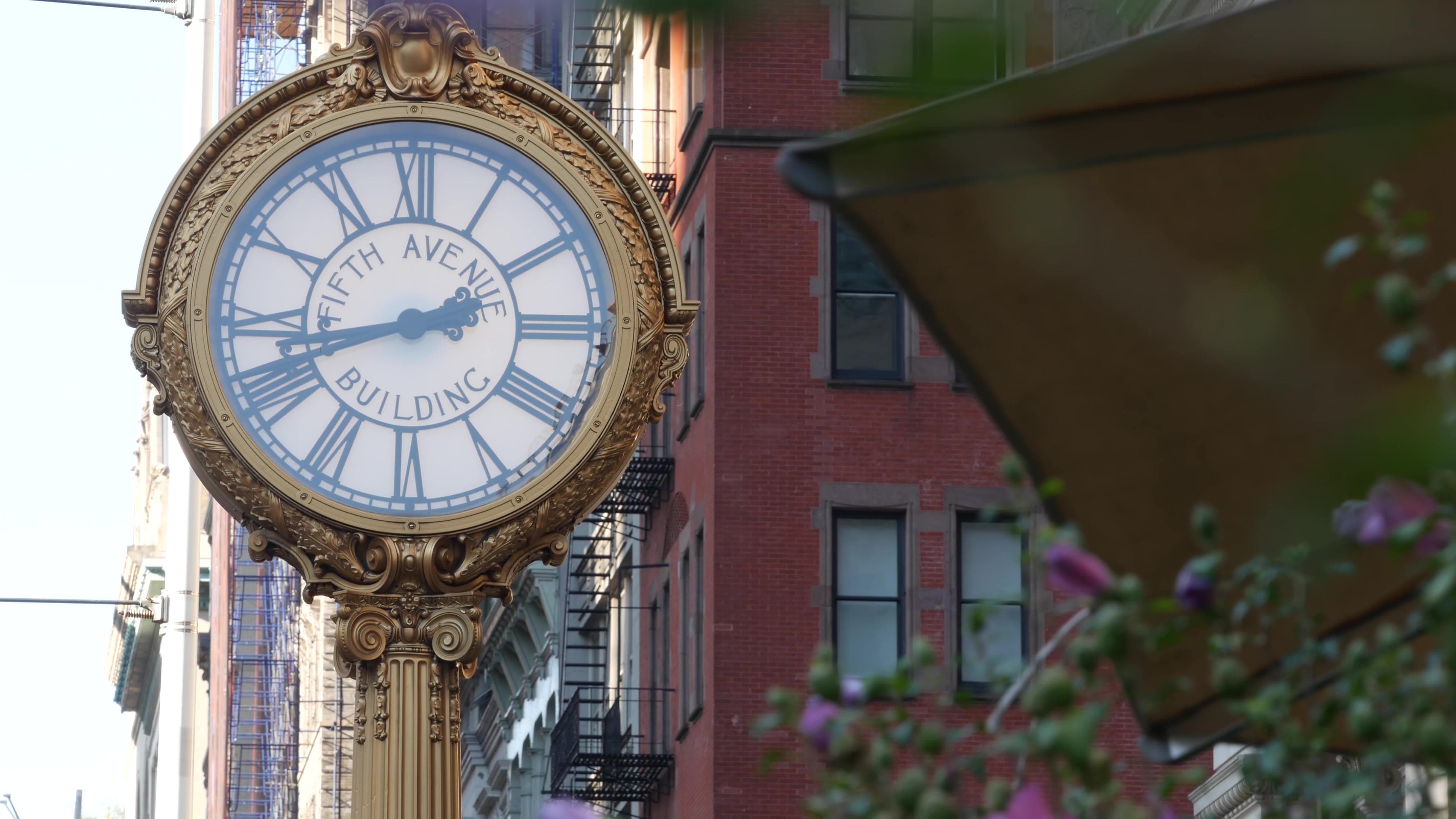 The gilded Fifth Avenue Building clock on a Flatiron District street, brick facades and an awning behind it