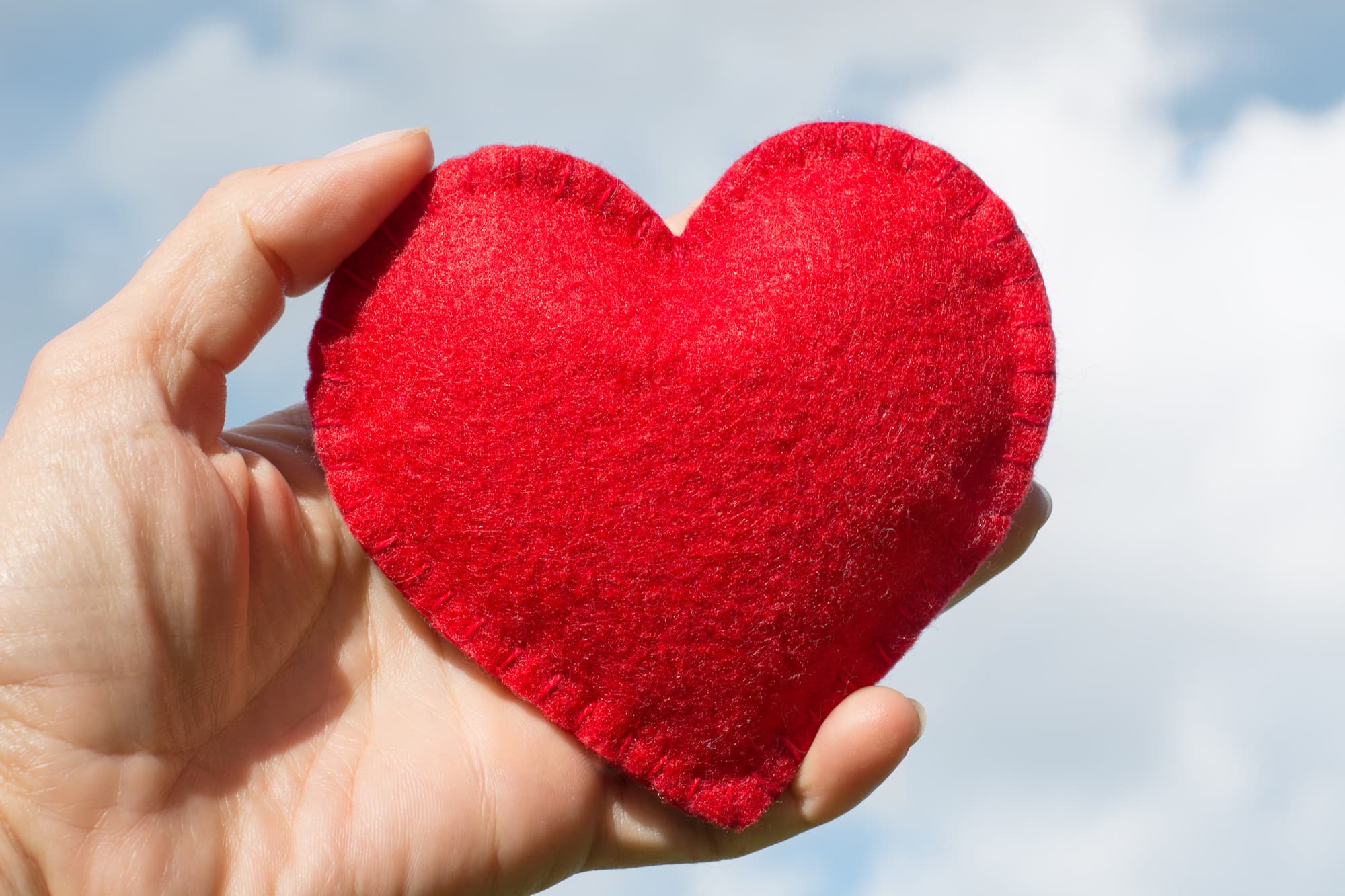 A hand holding a small red stitched heart up against a bright blue sky