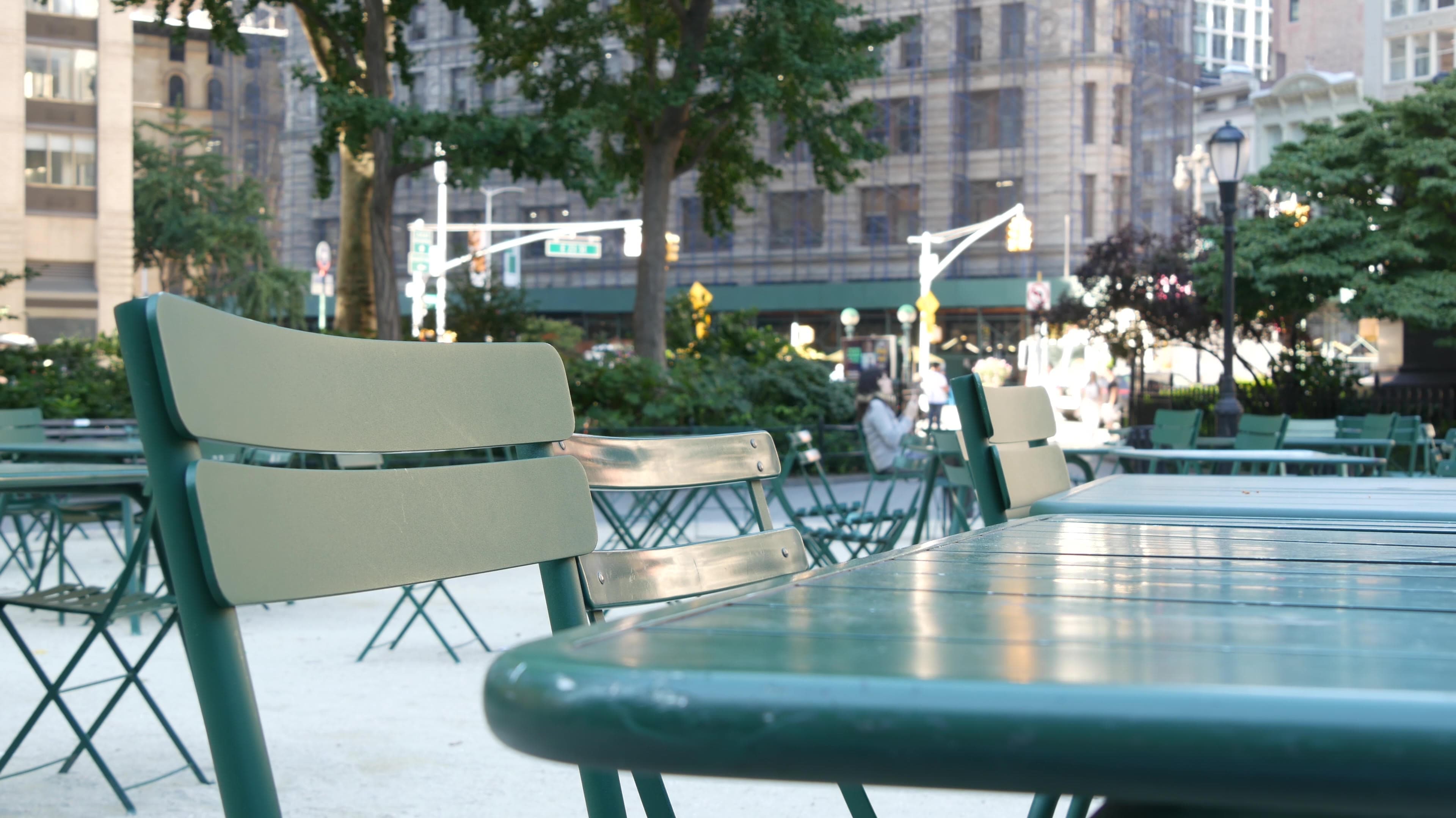 Green café tables and chairs in Madison Square Park, with the Flatiron District's buildings and trees soft behind them